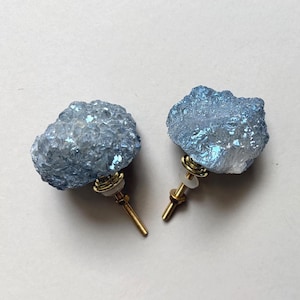 May include: Two cabinet knobs featuring raw, light blue crystalline clusters with a shimmering, iridescent finish. Each knob is attached to a gold-toned metal bolt with a threaded shaft, a small circular base, and a clear plastic washer for mounting.