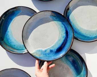 May include: A collection of ceramic plates with a unique design. Each plate features a light gray center with a vibrant blue and turquoise wave pattern around the rim. The plates have a dark brown edge.