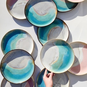 K&ouml;nnte beinhalten: Eine Sammlung von Keramiktellern mit einer blauen, gr&uuml;nen und wei&szlig;en Glasur. Die Teller haben ein einzigartiges, organisches Design mit einem wellenf&ouml;rmigen Muster. Einige der Teller haben eine rosa Glasur am Rand.
