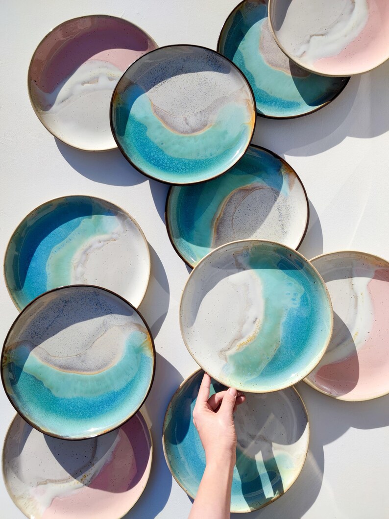 K&ouml;nnte beinhalten: Eine Sammlung von Keramiktellern mit einer blauen, gr&uuml;nen und wei&szlig;en Glasur. Die Teller haben ein einzigartiges, organisches Design mit einem wellenf&ouml;rmigen Muster. Einige der Teller haben eine rosa Glasur am Rand.