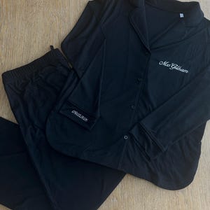 May include: Black pajama set with a button-down top and drawstring pants. The top features a notched collar and a breast pocket with the embroidered date "09.05.2028". The name "Mrs. Gillham" is embroidered on the front of the top in white.