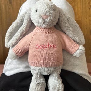 May include: A soft grey bunny toy with a pink knitted sweater. The sweater is embroidered with the name "Sophie". The bunny has long ears and a pink nose. The toy is designed for children.
