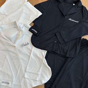 Puede incluir: Dos conjuntos de pijama, uno blanco y otro negro. El conjunto blanco tiene la palabra "Bride" bordada en la camisa y la fecha "3.05.2026" en el puño. El conjunto negro tiene la palabra "Bridesmaid" bordada en la camisa y la fecha "3.05.2026" en el puño.