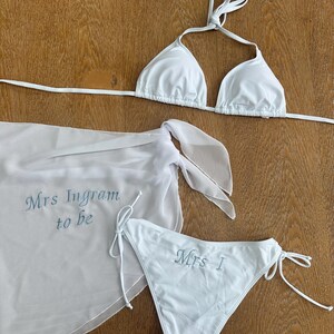 May include: A white bikini set consisting of a triangle top and side-tie bottoms, paired with a matching sheer sarong. The sarong features the embroidered text "Mrs Ingram to be" in blue, while the bikini bottom displays "Mrs I" in blue.