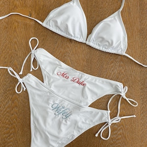 May include: A white bikini set consisting of a triangle top and two side-tie bikini bottoms. One bottom features the text Mrs Duke embroidered in red, while the other bottom features the text Wifey embroidered in a light blue script font.