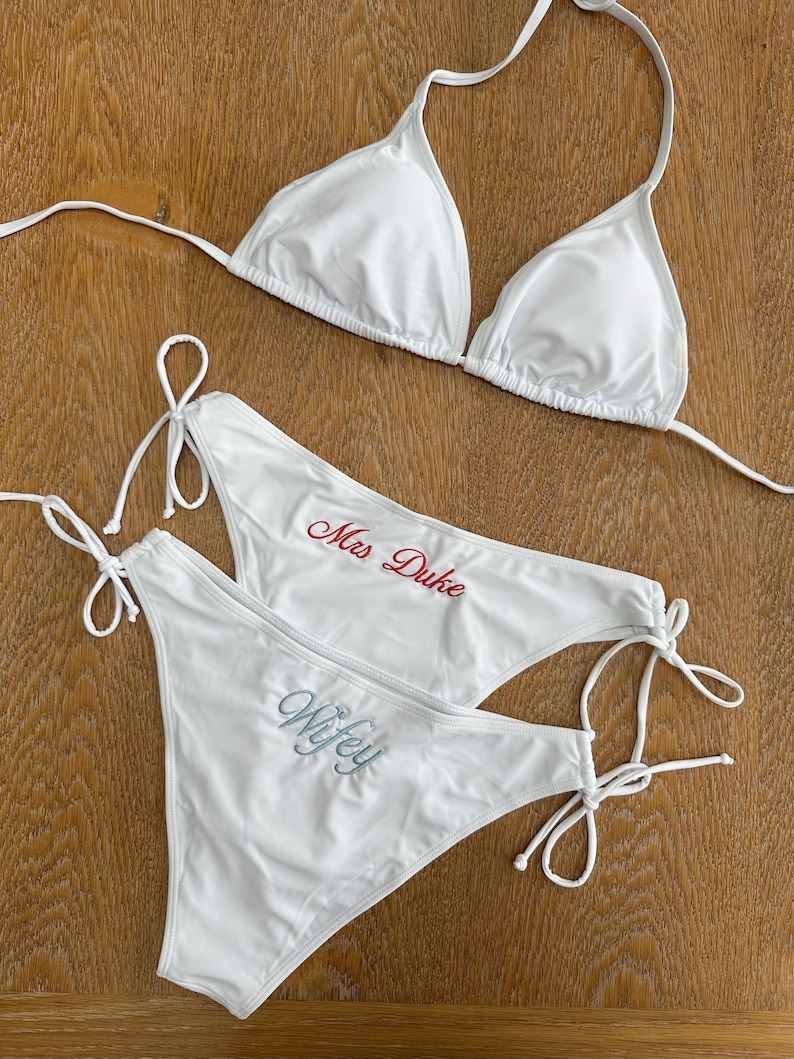 May include: A white bikini set consisting of a triangle top and two side-tie bikini bottoms. One bottom features the text Mrs Duke embroidered in red, while the other bottom features the text Wifey embroidered in a light blue script font.