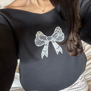 May include: Black sweatshirt with a white lace bow design. The sweatshirt is paired with white shorts with a red heart pattern.
