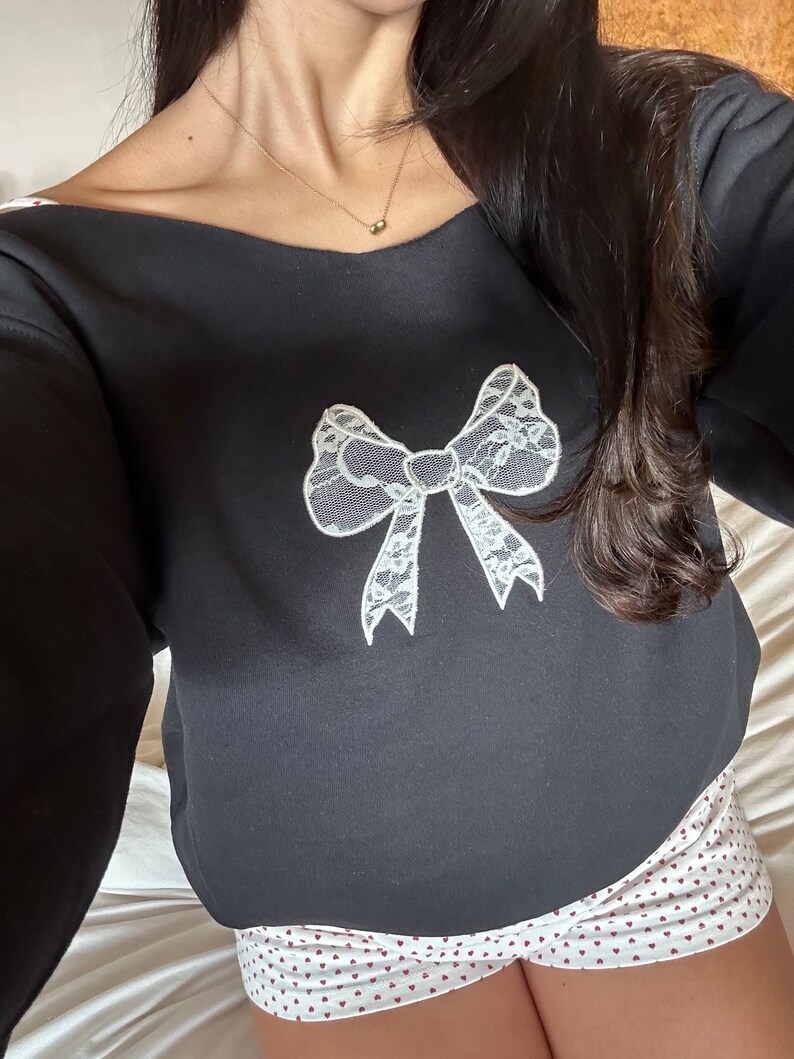 May include: Black sweatshirt with a white lace bow design. The sweatshirt is paired with white shorts with a red heart pattern.
