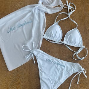 May include: A white bikini set consisting of a triangle top and side-tie bottoms, accompanied by a matching white sarong. Both the sarong and the bikini bottoms feature the embroidered text "Mrs Stephens" in a light blue cursive script.