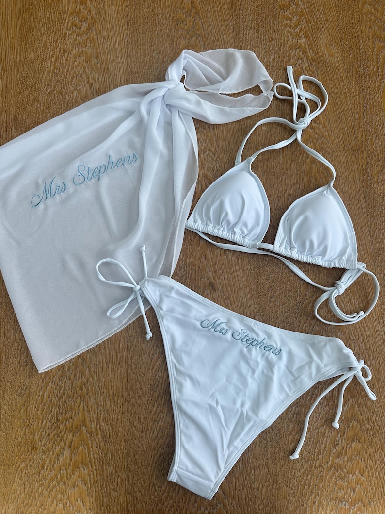 May include: A white bikini set consisting of a triangle top and side-tie bottoms, accompanied by a matching white sarong. Both the sarong and the bikini bottoms feature the embroidered text "Mrs Stephens" in a light blue cursive script.