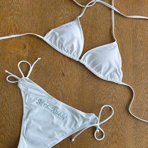 May include: A white two-piece bikini set featuring a triangle top with thin halter straps and matching side-tie bikini bottoms. The bottoms are embroidered with the text Mrs Russo in a light blue script font on the front left side.