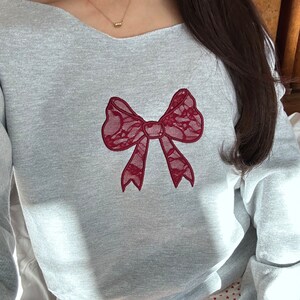 May include: A gray sweatshirt with a red lace bow embroidered on the front. The sweatshirt has a wide neckline and long sleeves.