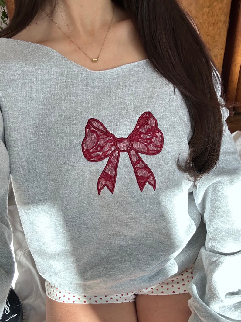 May include: A gray sweatshirt with a red lace bow embroidered on the front. The sweatshirt has a wide neckline and long sleeves.