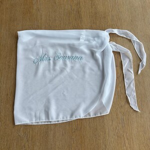 May include: A white sheer wrap skirt featuring the embroidered text Mrs. Sousana in light blue script. The garment is made of a lightweight, semi-transparent fabric and includes long, thin fabric ties on the side for securing the wrap around the waist.