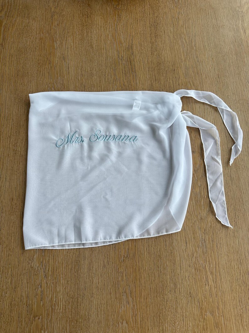 May include: A white sheer wrap skirt featuring the embroidered text Mrs. Sousana in light blue script. The garment is made of a lightweight, semi-transparent fabric and includes long, thin fabric ties on the side for securing the wrap around the waist.