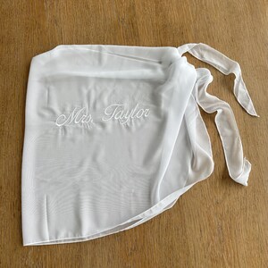May include: A white sheer wrap skirt featuring the embroidered text Mrs. Taylor in a cursive script font. The garment is constructed from a lightweight, semi-transparent fabric and includes long fabric ties on the side for securing the wrap.