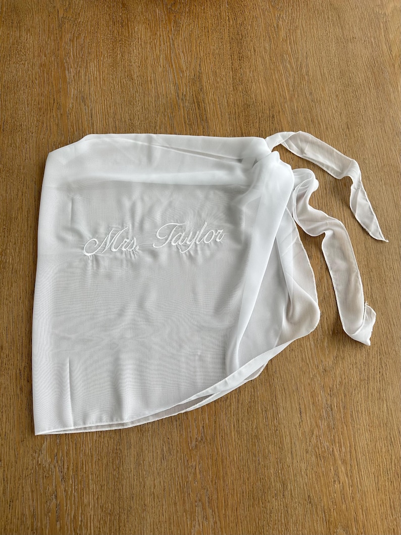 May include: A white sheer wrap skirt featuring the embroidered text Mrs. Taylor in a cursive script font. The garment is constructed from a lightweight, semi-transparent fabric and includes long fabric ties on the side for securing the wrap.