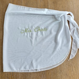 May include: A white sheer wrap skirt featuring the embroidered text Mrs. Chato in light green cursive script. The garment is made of a lightweight, semi-transparent fabric and includes integrated fabric ties on the side for a secure closure.