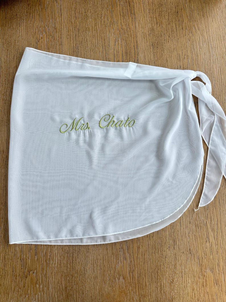 May include: A white sheer wrap skirt featuring the embroidered text Mrs. Chato in light green cursive script. The garment is made of a lightweight, semi-transparent fabric and includes integrated fabric ties on the side for a secure closure.
