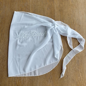 May include: A white sheer wrap skirt featuring the embroidered text "Mrs Beech" in a cursive script font. The garment is made of a lightweight, semi-transparent fabric and includes long fabric ties on the side for securing the wrap around the waist.