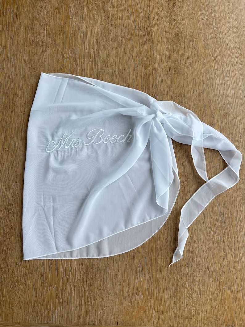 May include: A white sheer wrap skirt featuring the embroidered text "Mrs Beech" in a cursive script font. The garment is made of a lightweight, semi-transparent fabric and includes long fabric ties on the side for securing the wrap around the waist.