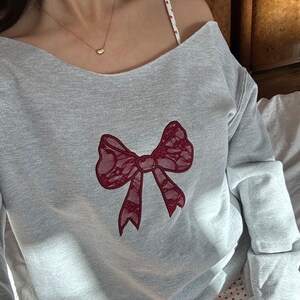May include: A gray sweatshirt with a red lace bow design. The sweatshirt has a scoop neck and long sleeves.