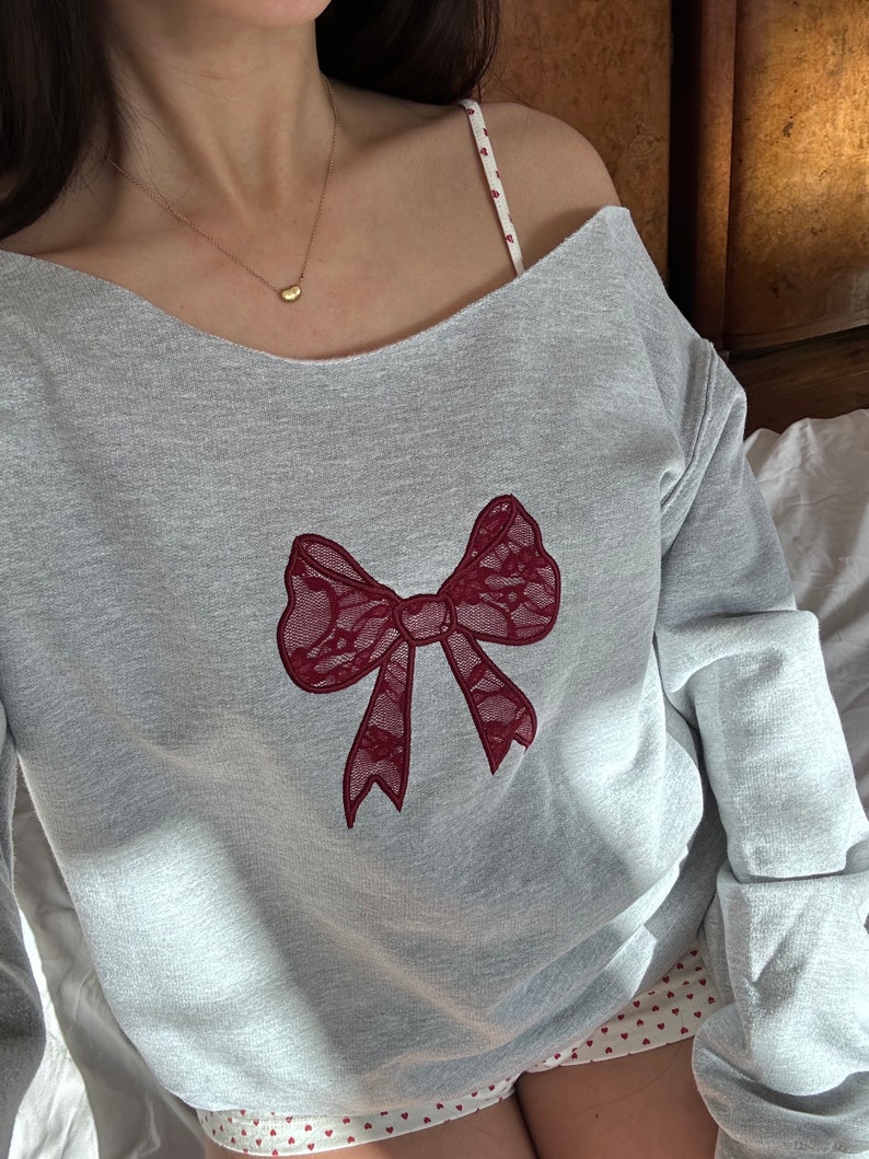 May include: A gray sweatshirt with a red lace bow design. The sweatshirt has a scoop neck and long sleeves.