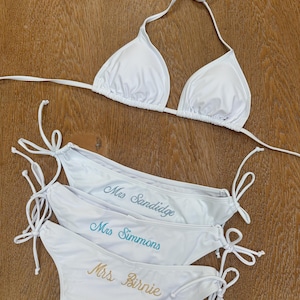 May include: A white bikini set consisting of a triangle top with halter straps and three matching bikini bottoms with side ties. Each bottom features custom embroidery in cursive script reading Mrs Sandidge, Mrs Simmons, and Mrs Birnie.