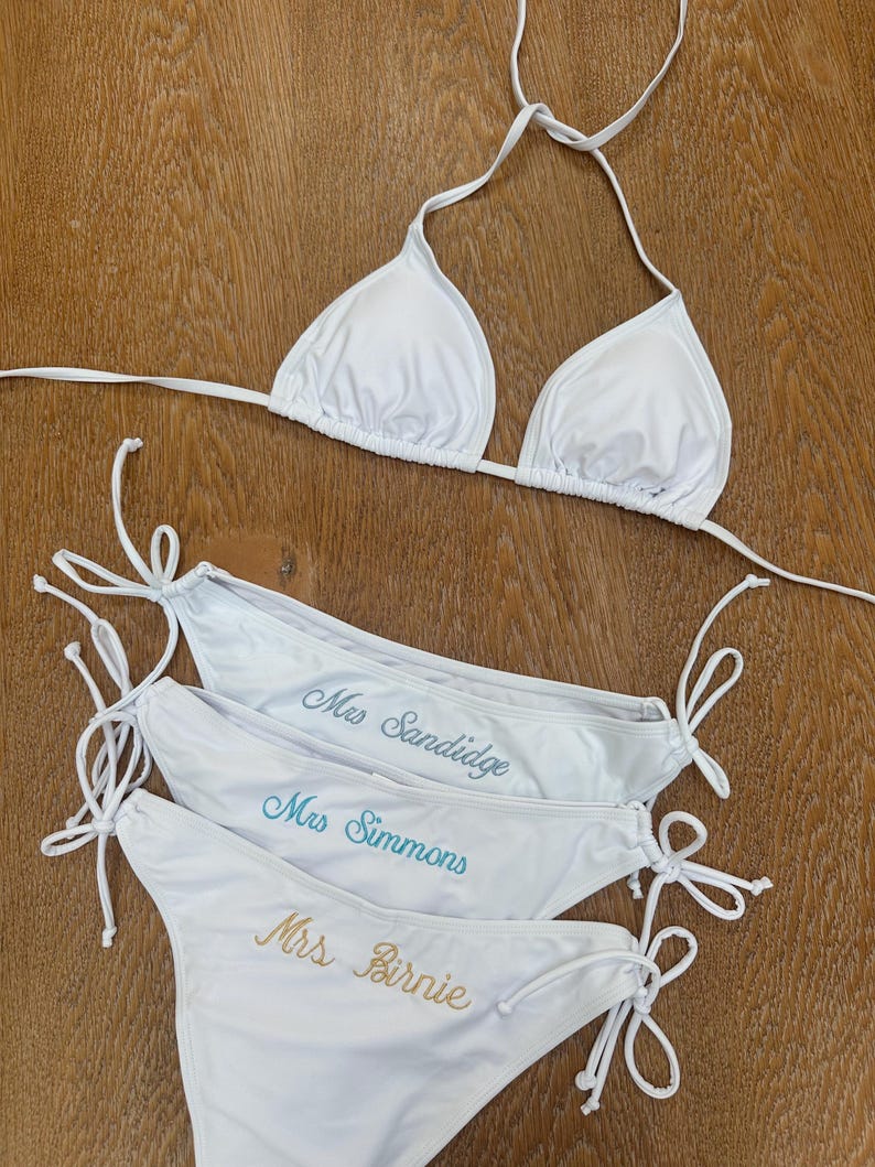 May include: A white bikini set consisting of a triangle top with halter straps and three matching bikini bottoms with side ties. Each bottom features custom embroidery in cursive script reading Mrs Sandidge, Mrs Simmons, and Mrs Birnie.