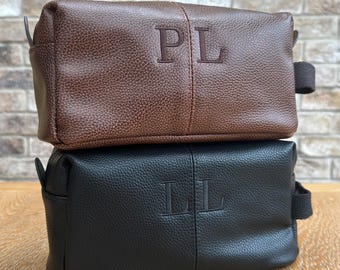May include: Two leather toiletry bags, one brown and one black, stacked on top of each other. The brown bag has the initials "PL" embossed on the front, while the black bag has "LL". Both bags have a rectangular shape.