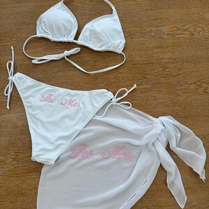 May include: A white bikini set consisting of a triangle top and side-tie bottoms, paired with a matching sheer white sarong. The bikini bottoms and the sarong both feature the text "The Mrs." embroidered in light pink script lettering.