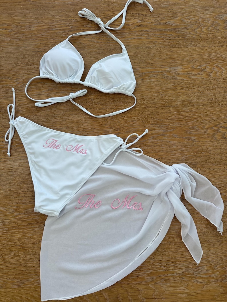 May include: A white bikini set consisting of a triangle top and side-tie bottoms, paired with a matching sheer white sarong. The bikini bottoms and the sarong both feature the text "The Mrs." embroidered in light pink script lettering.
