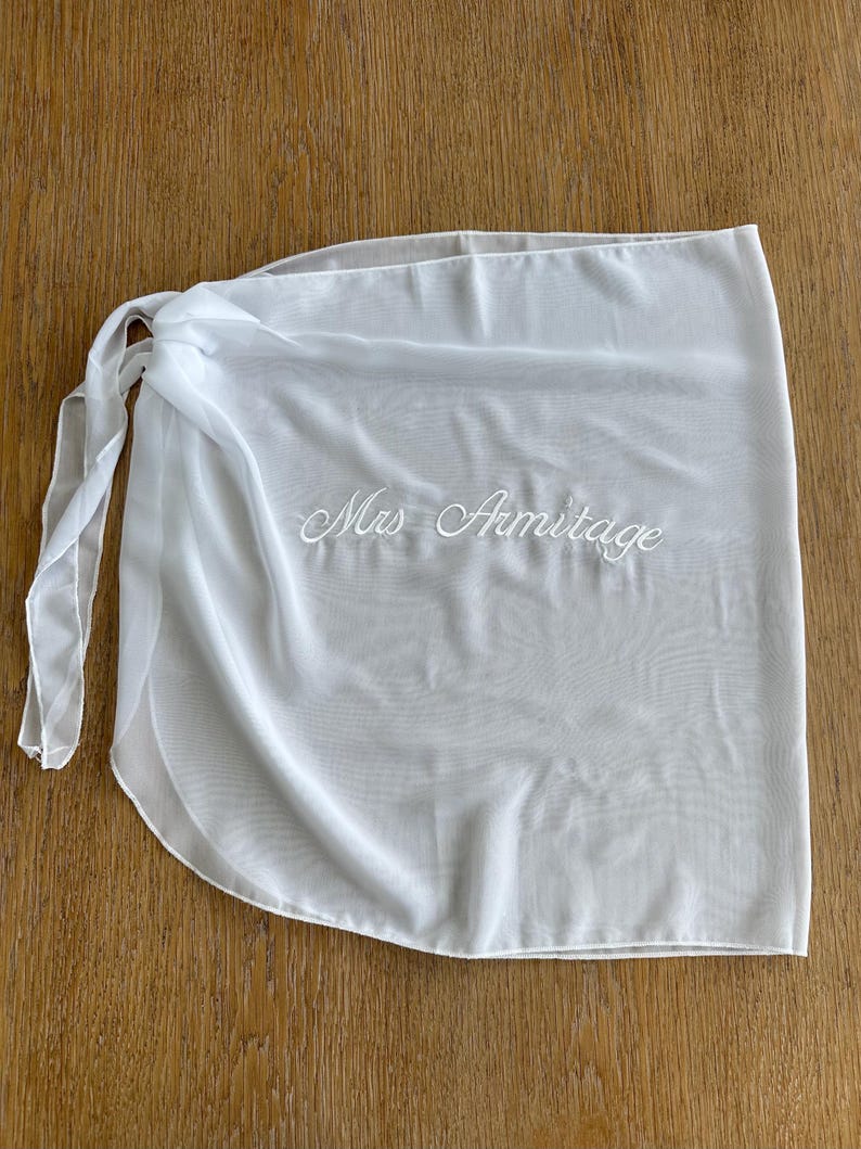May include: A white sheer wrap skirt featuring a tie closure on the left side. The fabric is lightweight and semi-transparent. Embroidered in white cursive script across the center of the skirt is the text Mrs Armitage, clearly visible.
