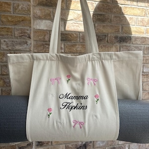 May include: A white canvas tote bag with a gray yoga mat inside. The bag has pink and white embroidered bows and roses around the edge. The text "Mamma Hopkins" is embroidered in black in the center of the bag.