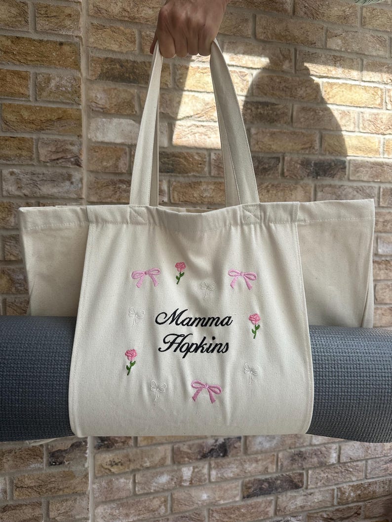 May include: A white canvas tote bag with a gray yoga mat inside. The bag has pink and white embroidered bows and roses around the edge. The text "Mamma Hopkins" is embroidered in black in the center of the bag.