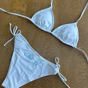 May include: A white two-piece bikini set featuring a triangle top with thin tie straps and matching side-tie bottoms. The bikini bottoms are embroidered with the text "Mrs E" in a light blue cursive script font on the front left side.