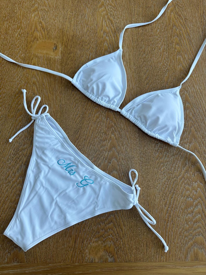 May include: A white two-piece bikini set featuring a triangle top with thin tie straps and matching side-tie bottoms. The bikini bottoms are embroidered with the text "Mrs E" in a light blue cursive script font on the front left side.