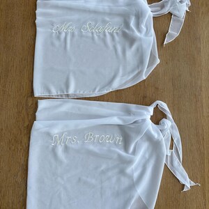 May include: Two white sheer wrap skirts featuring tie closures at the side. The top skirt is embroidered with the text Mrs. Sclafani in white script, and the bottom skirt is embroidered with the text Mrs. Brown in white script.