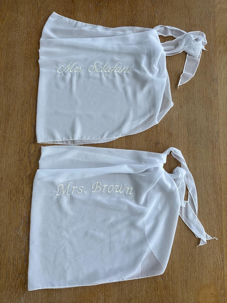 May include: Two white sheer wrap skirts featuring tie closures at the side. The top skirt is embroidered with the text Mrs. Sclafani in white script, and the bottom skirt is embroidered with the text Mrs. Brown in white script.