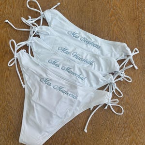 May include: Four white bikini bottoms with side ties, each featuring custom embroidery in a cursive script. The names embroidered on the fabric are Mrs. Stephens, Mrs. Wanstall, Mrs. Marechal, and Mrs. Mancera, displayed in a stacked arrangement.