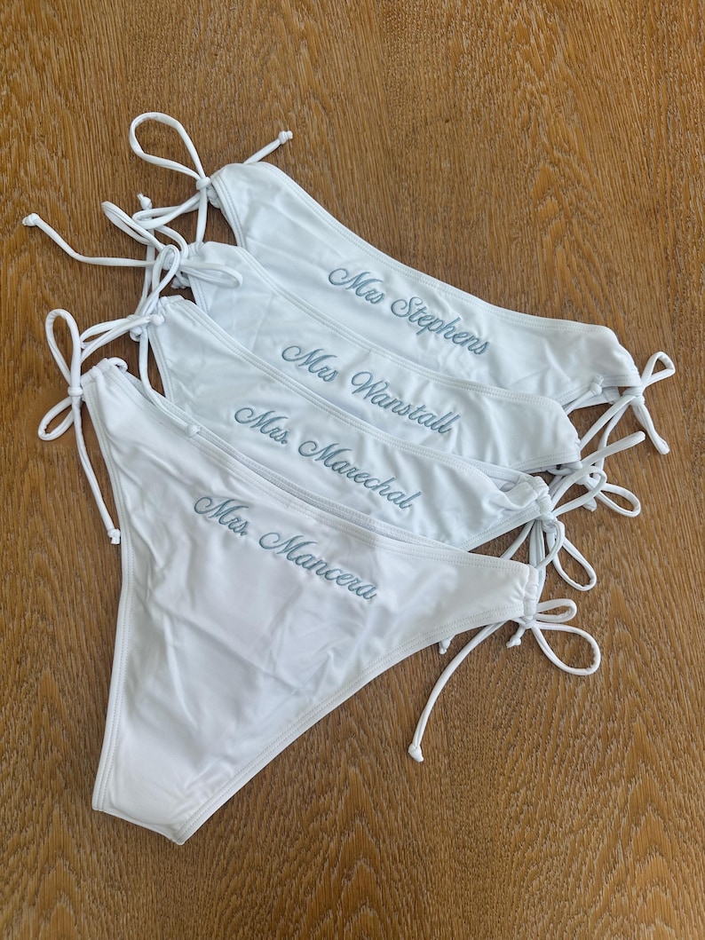 May include: Four white bikini bottoms with side ties, each featuring custom embroidery in a cursive script. The names embroidered on the fabric are Mrs. Stephens, Mrs. Wanstall, Mrs. Marechal, and Mrs. Mancera, displayed in a stacked arrangement.