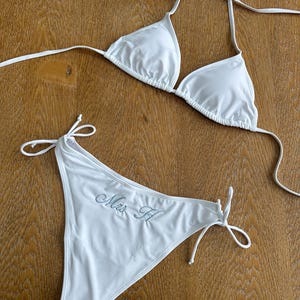 May include: A white two-piece bikini set featuring a triangle top with halter neck ties and matching bikini bottoms with side ties. The front of the bikini bottoms features the embroidered text "Mrs H" in a light blue cursive script font.