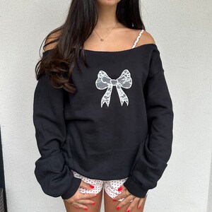 May include: Black off-the-shoulder sweatshirt with a white lace bow design.