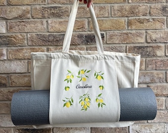 May include: A cream-colored tote bag with a gray yoga mat. The tote features an embroidered lemon design and the name "Carolina." The yoga mat is rolled up and secured by the tote. The bag is held up against a brick wall.
