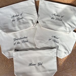 May include: Five off-white canvas zippered pouches featuring black embroidered text. The pouches are labeled Mother of the Groom, Mother of the Bride, Grandmother of the Bride, Maid of Honor, and Flower Girl in a cursive script font.