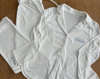 Puede incluir: Conjunto de pijama blanco con mangas largas y pantalón. La camisa tiene una parte delantera abotonada y la palabra "Bride" bordada en azul claro. El puño de la manga tiene la fecha "8.08.26" bordada en azul claro.
