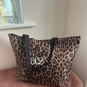 Puede incluir: Bolso tote con estampado de leopardo en tonos marrón y negro. El bolso tiene asas negras y las iniciales blancas "SL". El bolso está sobre una superficie rosa. Adecuado para llevar objetos.