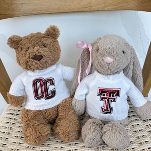 Könnte beinhalten: Ein brauner Plüschteddybär und ein hellbrauner Plüschhase sitzen nebeneinander. Beide Stofftiere tragen weiße Strickpullover. Der Pullover des Bären zeigt die Buchstaben OC in Rot und Schwarz, während der Pullover des Hasen ein rotes und schwarzes TT-Logo aufweist. Der Hase hat ein kleines rosa Band an einem Ohr.