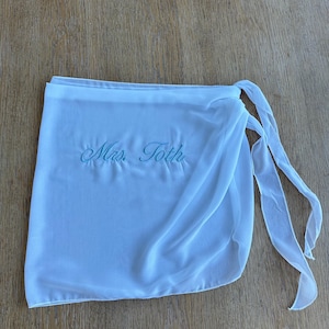 May include: A white wrap skirt made of sheer fabric featuring the embroidered text "Mrs. Toth" in light blue script. The garment includes a waistband and long fabric ties on the right side for securing the wrap around the waist.