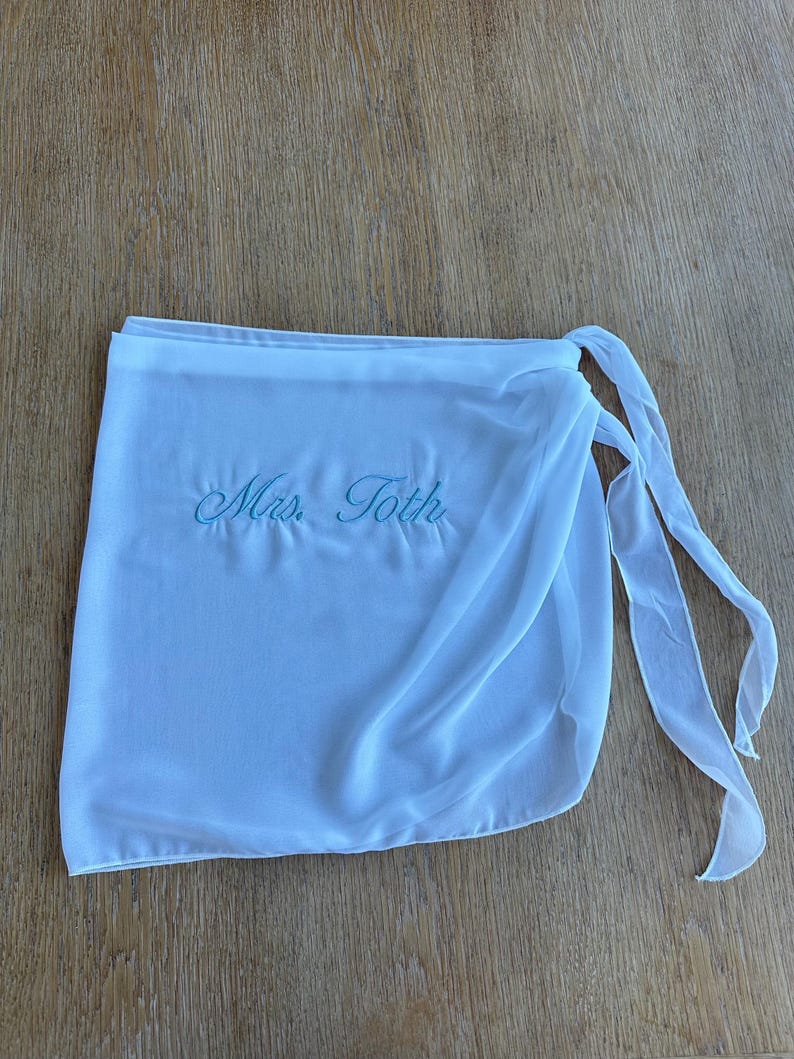 May include: A white wrap skirt made of sheer fabric featuring the embroidered text "Mrs. Toth" in light blue script. The garment includes a waistband and long fabric ties on the right side for securing the wrap around the waist.
